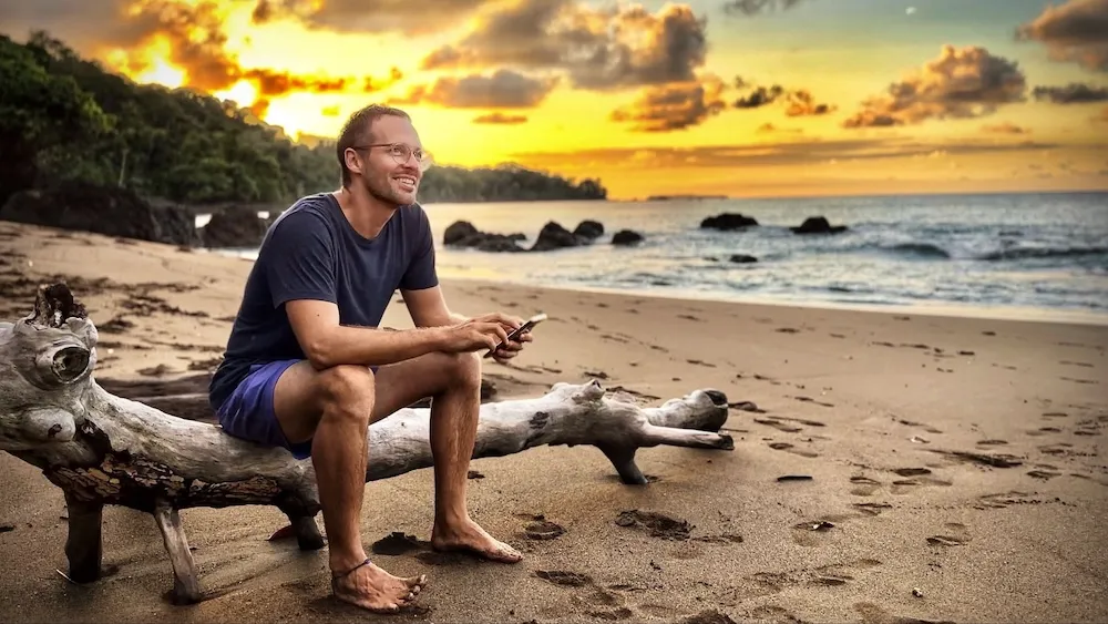 Ernst sitzt am Strand bei Sonnenuntergang mit dem Smartphone – Business läuft, während er das Leben genießt